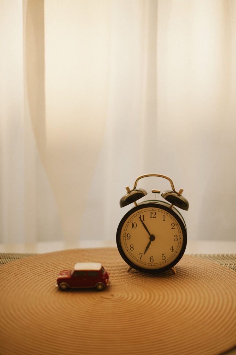A small red toy car posed next to an old-fashioned alarm clock on a beige surface.